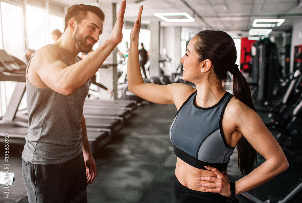 Man and woman in a gym high-fiving, smiling, with treadmills and fitness equipment in the background.